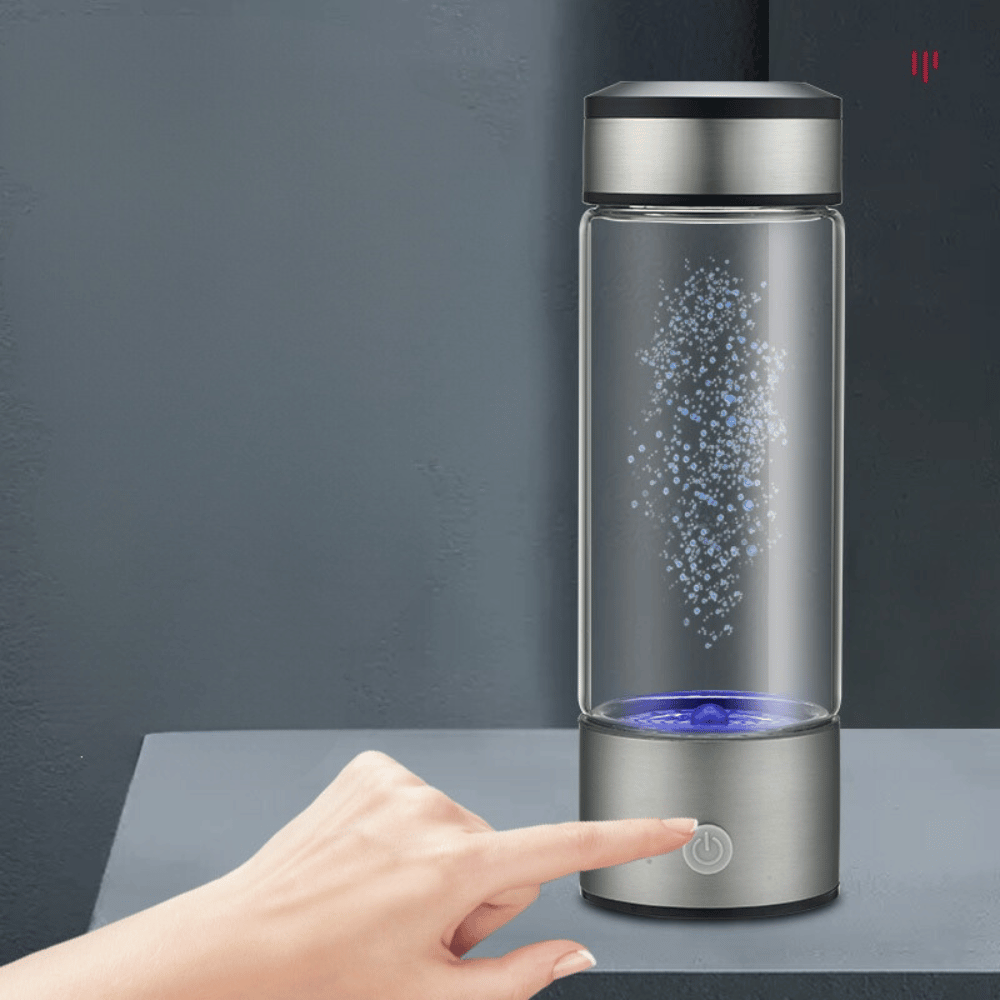 Glass water bottle with a silver base and black lid on a dark surface, with a hand pressing a button.