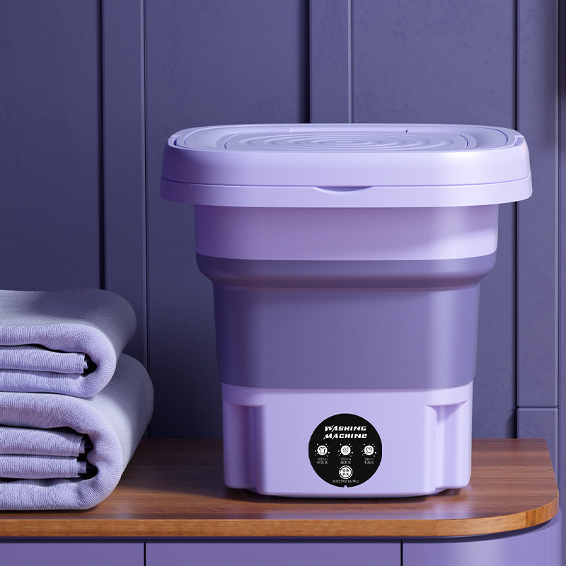Purple folding washing machine on a wooden surface with folded clothes against a purple wall.
